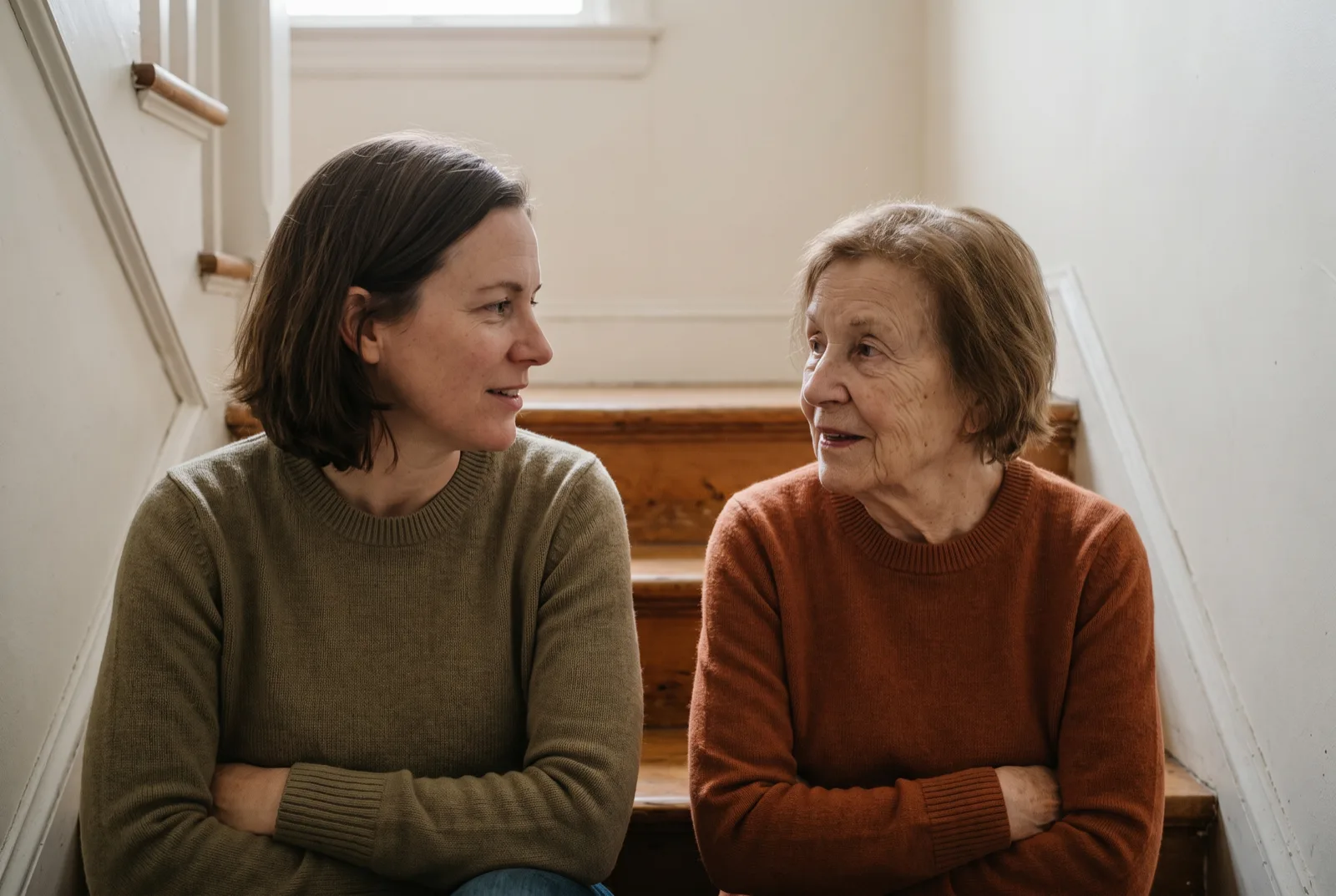 A woman seated on the staircase of an older home talking with her elderly mother a few steps above her